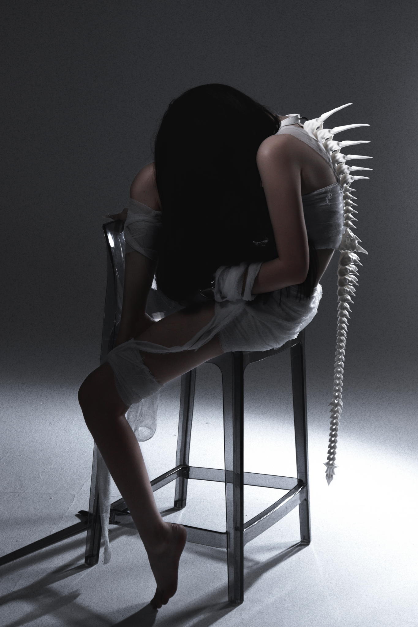 Studio portrait of a seated figure leaning forward, with white fabric and a sculptural spine accessory