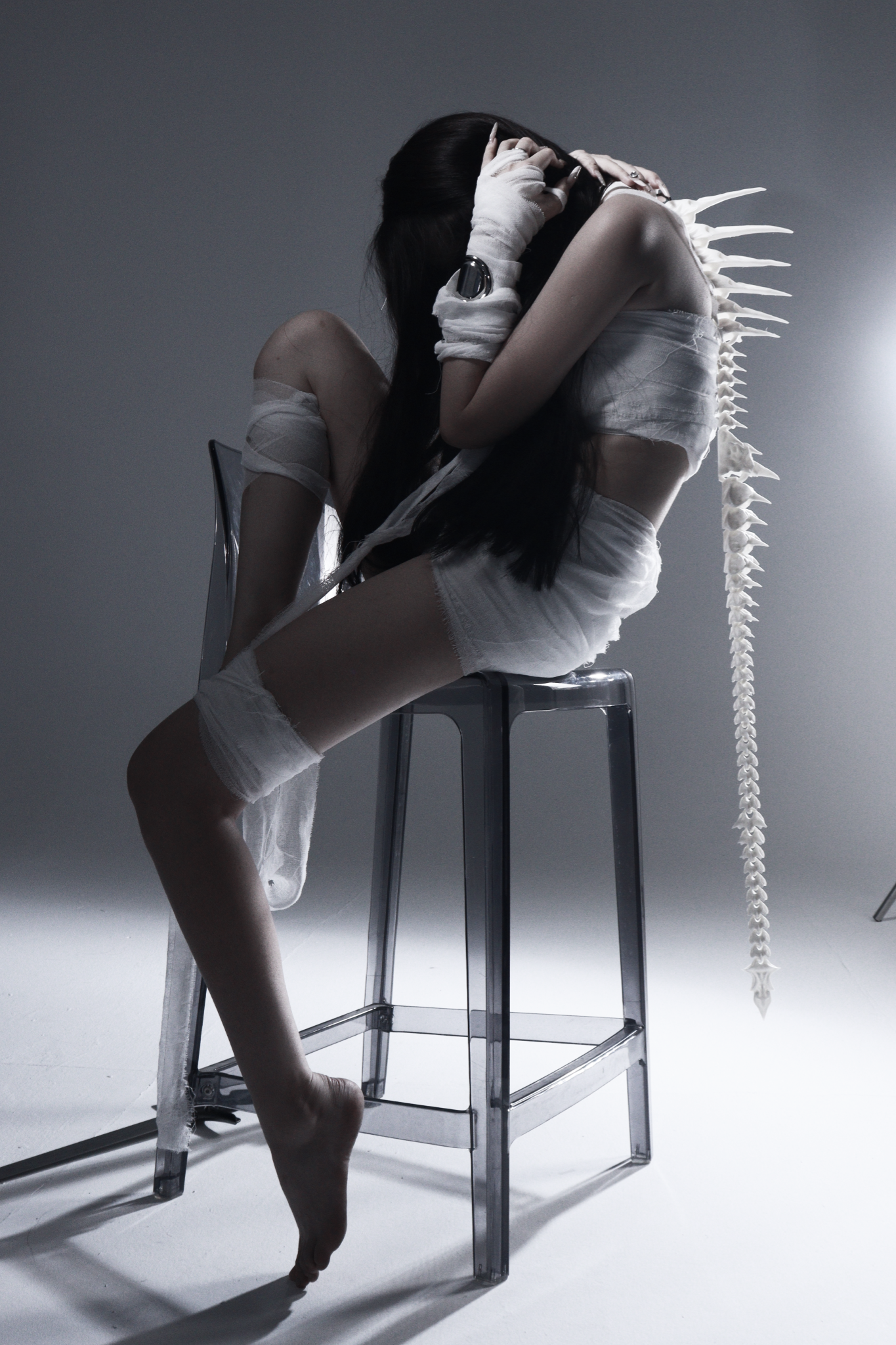 Studio portrait of a seated figure with hands raised to their head and a sculptural spine accessory